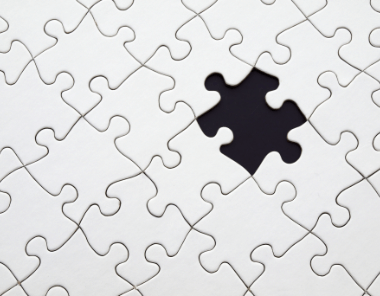 A close-up of a white jigsaw puzzle with one piece missing, revealing a dark empty space where the piece should fit.