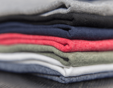 A close-up view of a neatly stacked pile of folded clothes in various colors, including gray, black, navy blue, red, green, white, and blue, on a dark surface.