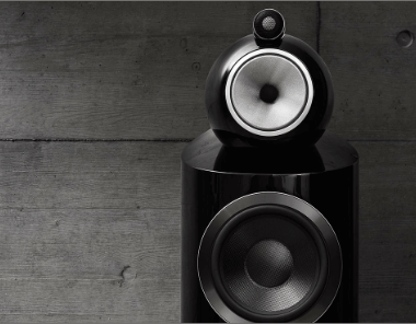 A close-up of a modern black speaker with two circular drivers, set against a textured gray concrete wall. The speaker has a sleek, glossy finish and a minimalist design.
