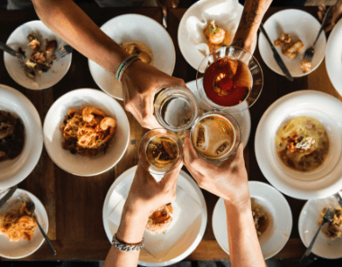 Four people toast with drinks over a wooden table set with plates of pasta, shrimp, and other food. The image is viewed from above, capturing a celebratory meal.
