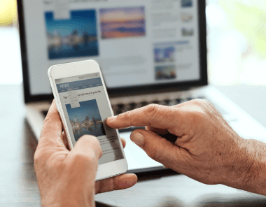 A person uses a smartphone to scroll through a news article, while an open laptop displaying a similar webpage is visible in the background.