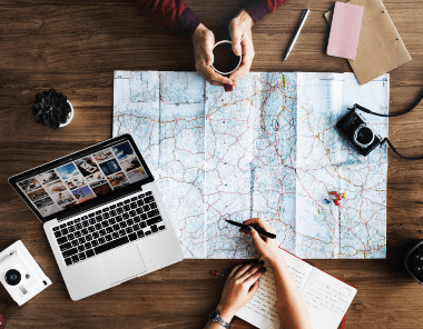 Two people plan a trip at a wooden table with a large map, notebook, laptop, camera, plant, cup of coffee, and travel photos spread out. One writes in a notebook while the other holds a mug.