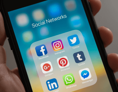 A hand holding a smartphone displaying a folder labeled Social Networks with icons for Facebook, Instagram, Twitter, Google+, Pinterest, Tumblr, LinkedIn, WhatsApp, and Messenger.