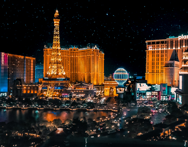 A vibrant night view of the Las Vegas Strip with brightly lit hotels, including a replica of the Eiffel Tower, glowing signs, and bustling city lights against a dark sky.