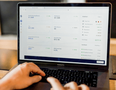 A person using a MacBook Pro is viewing a financial dashboard displaying revenue, net income, graphs, and breakdowns of business performance metrics.
