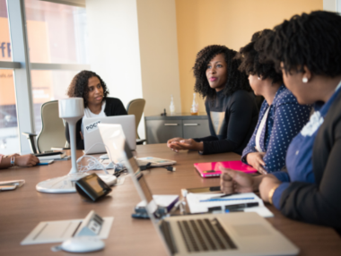 Five women sit around a conference table with laptops, notebooks, and paperwork, engaged in a discussion in a modern office setting with large windows and orange walls.