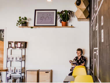 A person sits on a yellow chair in a modern cafe, reading a book. There are plants and framed art on a wall shelf above, with coffee products on nearby shelves and a chalkboard wall to the right.