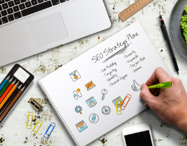 A person writes SEO Strategy Plan on a notepad surrounded by colorful icons. Nearby are a laptop, smartphone, ruler, salad, and scattered paper clips on a white desk.