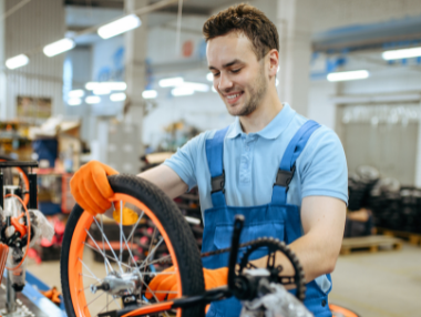 A young man in blue overalls and orange gloves smiles while assembling a bicycle wheel in a workshop, surrounded by tools and bike parts—dedicated to designing a digital experience for everyone.