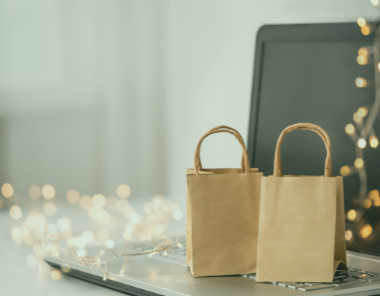 Two small brown paper shopping bags sit on a laptop keyboard with a blurred background of warm fairy lights, creating a festive mood that hints at online shopping and Tips to Prep Your eCommerce Store for the Holiday Season.