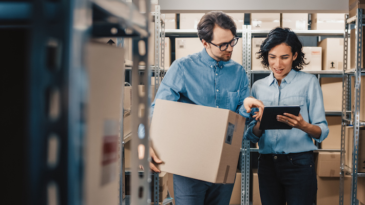 Two people stand in a warehouse surrounded by shelves with boxes. One person holds a large cardboard box while the other points at a tablet, both appearing to discuss B2B eCommerce strategies for inventory or logistics.
