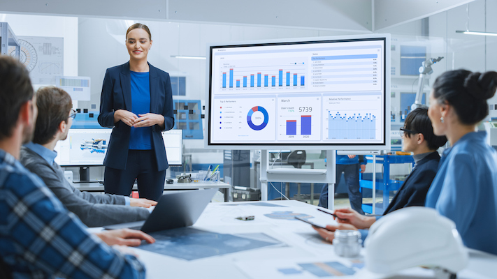 A woman stands and presents on How B2B Businesses Use Data, displaying graphs and analytics to colleagues in a modern office meeting room. Laptops and documents are spread across the table as the team analyzes key insights.