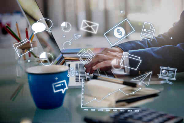 A person typing on a laptop at a desk with a blue coffee mug, papers, and animated business icons like charts, emails, and folders overlaid, symbolizing digital business or online communication.