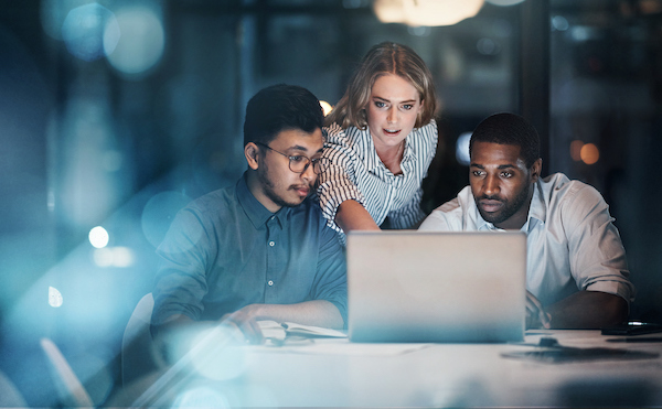 Three people work together at a desk, focused on a laptop screen in a dimly lit office. As they discuss, one woman stands and points at the screen, leading a collaborative effort to switch eCommerce platforms.