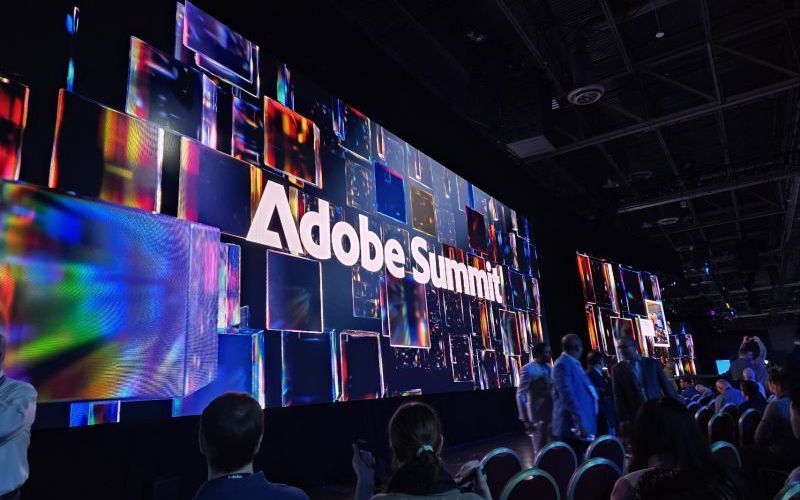 A large digital screen at an event displays the words Adobe Summit with abstract colorful graphics, as attendees sit and stand in a dimly lit auditorium awaiting key takeaways.