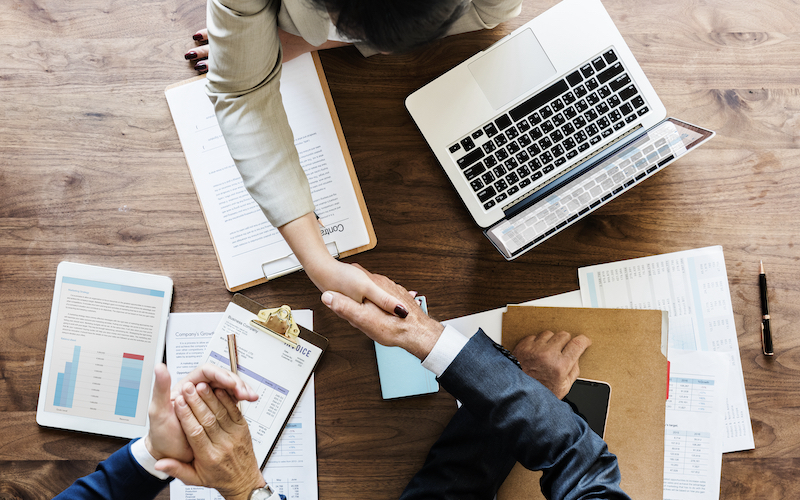 Two people in business attire shake hands across a wooden desk with a laptop, charts, documents, and a clipboard—signifying a partnership powered by eCommerce technology.