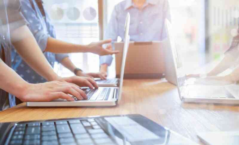 Several people standing around a table working on laptops, with hands typing on keyboards and sunlight streaming in. The scene suggests teamwork and collaboration in a B2B business office setting.