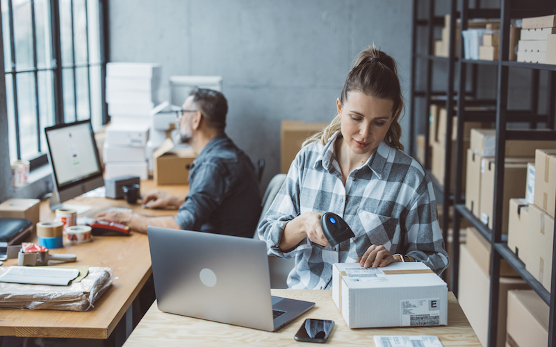 A woman scans a shipping label on a package at a desk with a laptop—perfect for B2B companies—while a man works on a computer in the background, surrounded by shelves filled with boxes.