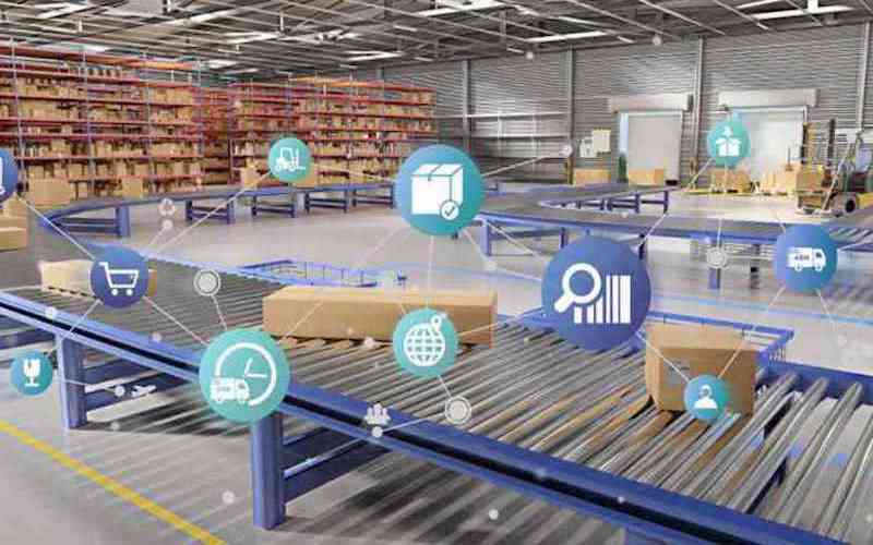A warehouse with conveyor belts, boxes, and shelves; digital icons overlay the image, representing data, shipping, inventory, shopping carts, trucks, and analytics—illustrating how manufacturers and distributors use smart logistics and supply chain technology.