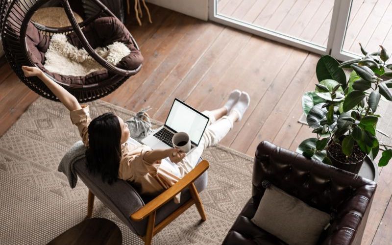 A person relaxes in an armchair with a laptop on their lap, stretching one arm and holding a mug in the other, enjoying the virtual work world. The room features wooden floors, a hanging chair, a plant, and large windows.