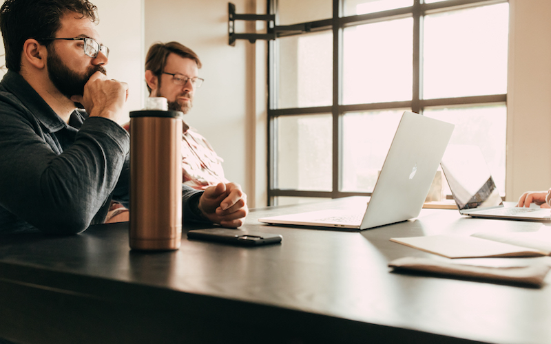 Two men with beards sit at a table in front of laptops, appearing focused as they discuss strategies for their B2B ecommerce site. A metallic travel mug and a smartphone rest on the table, while sunlight streams through large windows in the background.