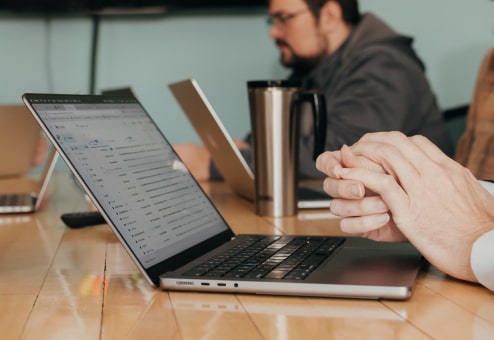 A person sits at a table with hands clasped in front of an open laptop displaying an email inbox, while another person in the background analyzes data and reviews analytics on their own laptop.