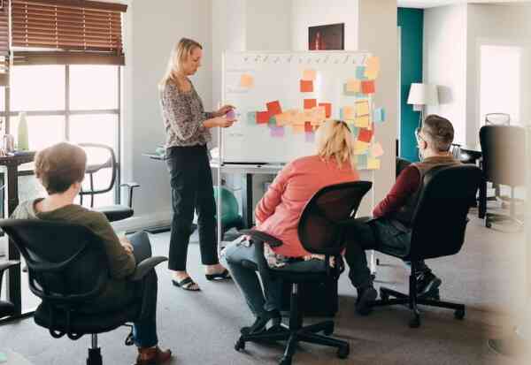 Four people in an office sit around a whiteboard covered with colorful sticky notes, while one person stands and presents, holding a marker. The room has large windows and modern office chairs.