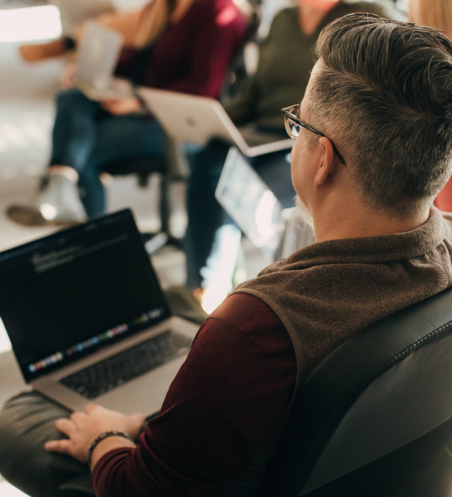 A person with short hair, wearing glasses and a brown shirt, sits in a chair working on a laptop. Others with laptops are seated in a circle, suggesting a group discussion or meeting in a well-lit room.