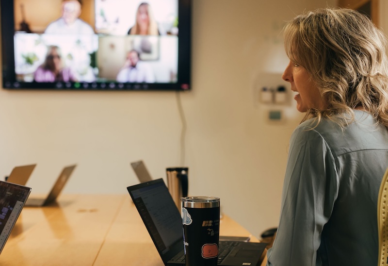 A person sitting at a conference table with a laptop, participating in a video conference. A large screen displays four people in a video call. A tumbler and another laptop are also on the table.