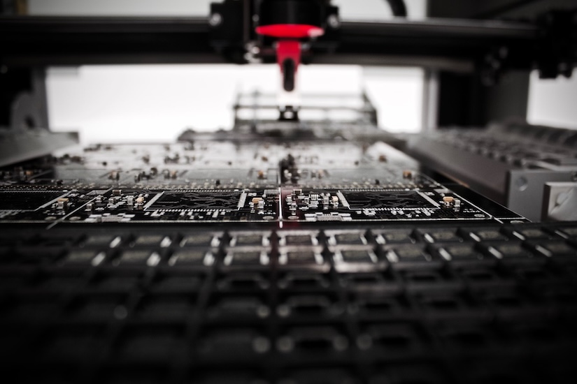 Close-up of a printed circuit board (PCB) manufacturing process. The image shows intricate electronic components and circuits on a PCB under a red laser, suggesting precision engineering and technology.