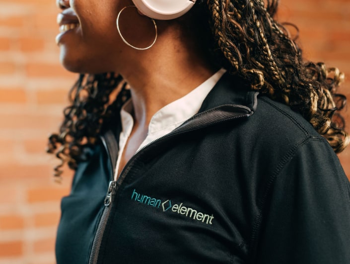 A woman with curly hair wearing pink headphones and hoop earrings smiles. She is dressed in a black jacket with the words human element embroidered on it. A brick wall serves as the backdrop.