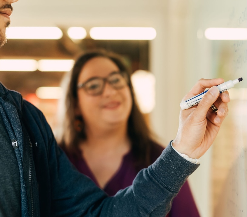 A person is writing on a whiteboard with a marker while another person stands nearby, smiling. The background is softly blurred, indicating an indoor setting with bright lighting.
