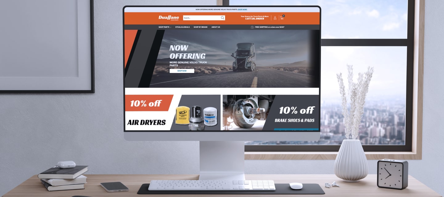 Computer monitor displaying a website with automotive products, including air dryers and brake shoes on discount. The background shows a road with a truck. The desk has a keyboard, mouse, plant, cup, and clock, with a window view.