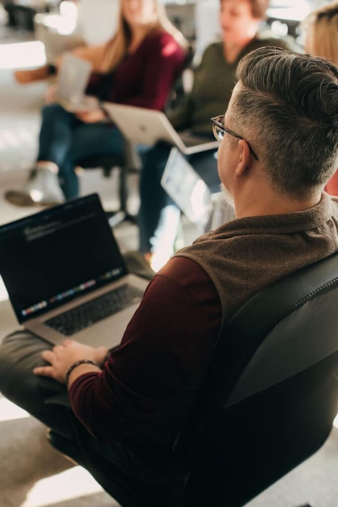 A group of people sit in a circle during a casual meeting. One person in the foreground, with glasses and a laptop, is turned towards others. Two people with open laptops are visible in soft focus in the background.
