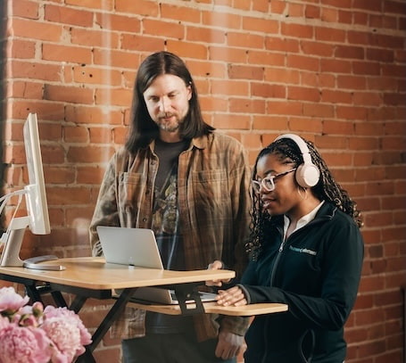 Two people are working at a standing desk in front of a brick wall. One person is typing on a laptop while wearing headphones, and the other is looking at the screen. There is a vase of pink flowers on the desk.